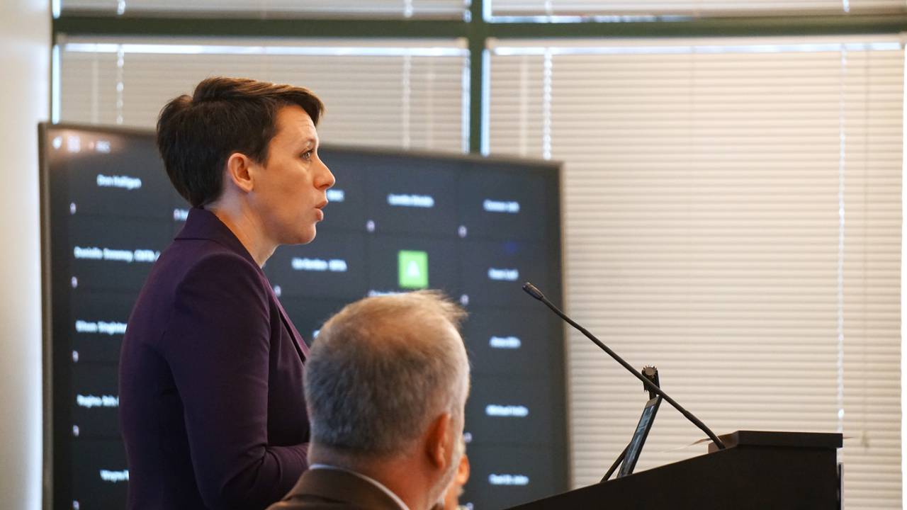 A close up photo of a woman standing and speaking at a podium while a television in the background displays virtual meeting attendees on platform Zoom.