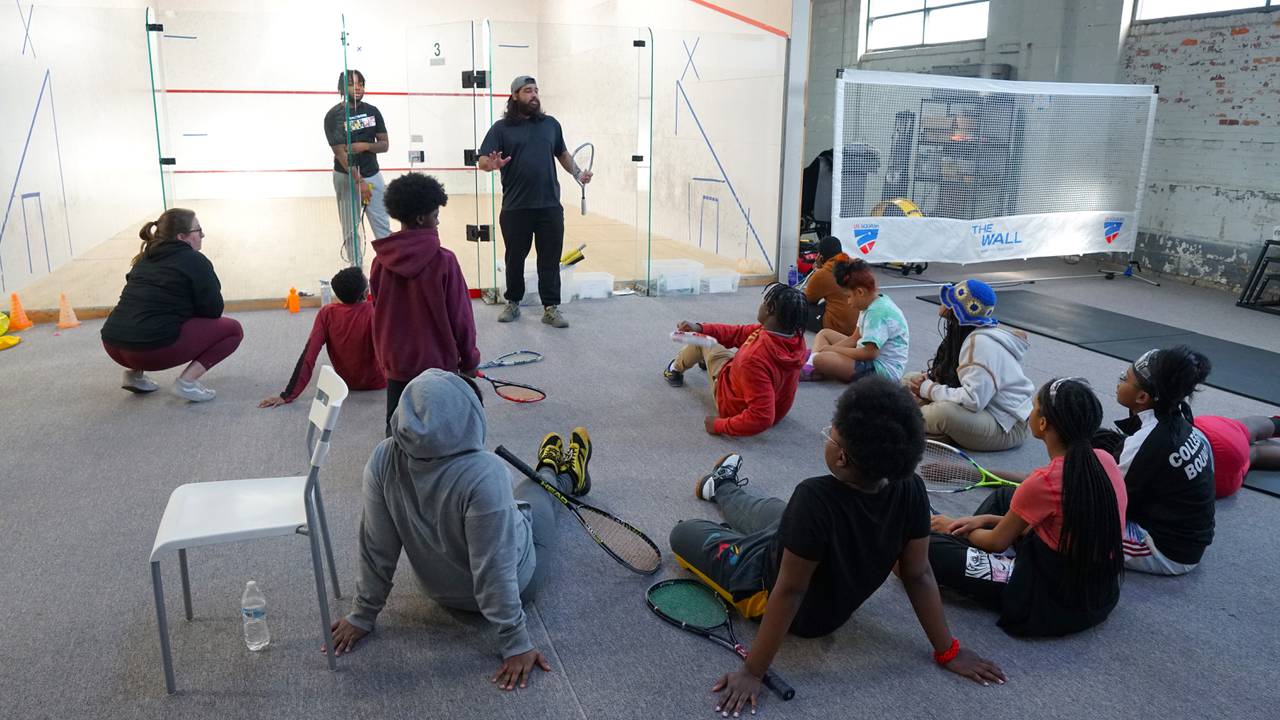 Two adults stand between a white squash court and a group of middle school students who are sitting on the ground watching them and holding squash rackets.