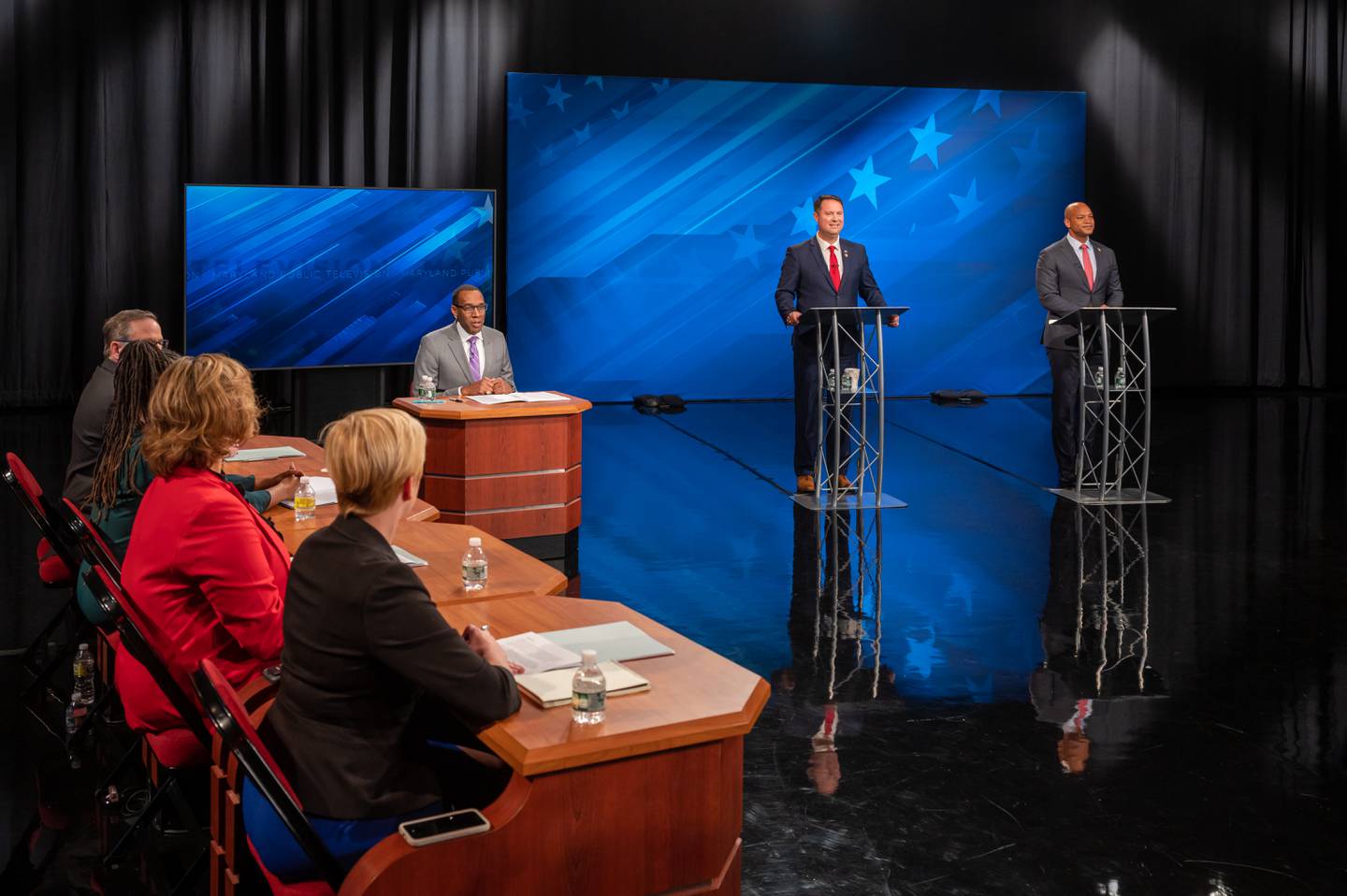 Dan Cox stands at a lectern in the center of the stage. Wes Moore stands at a lectern stage left. Stage right, the debate moderator and panelists sit at desks.