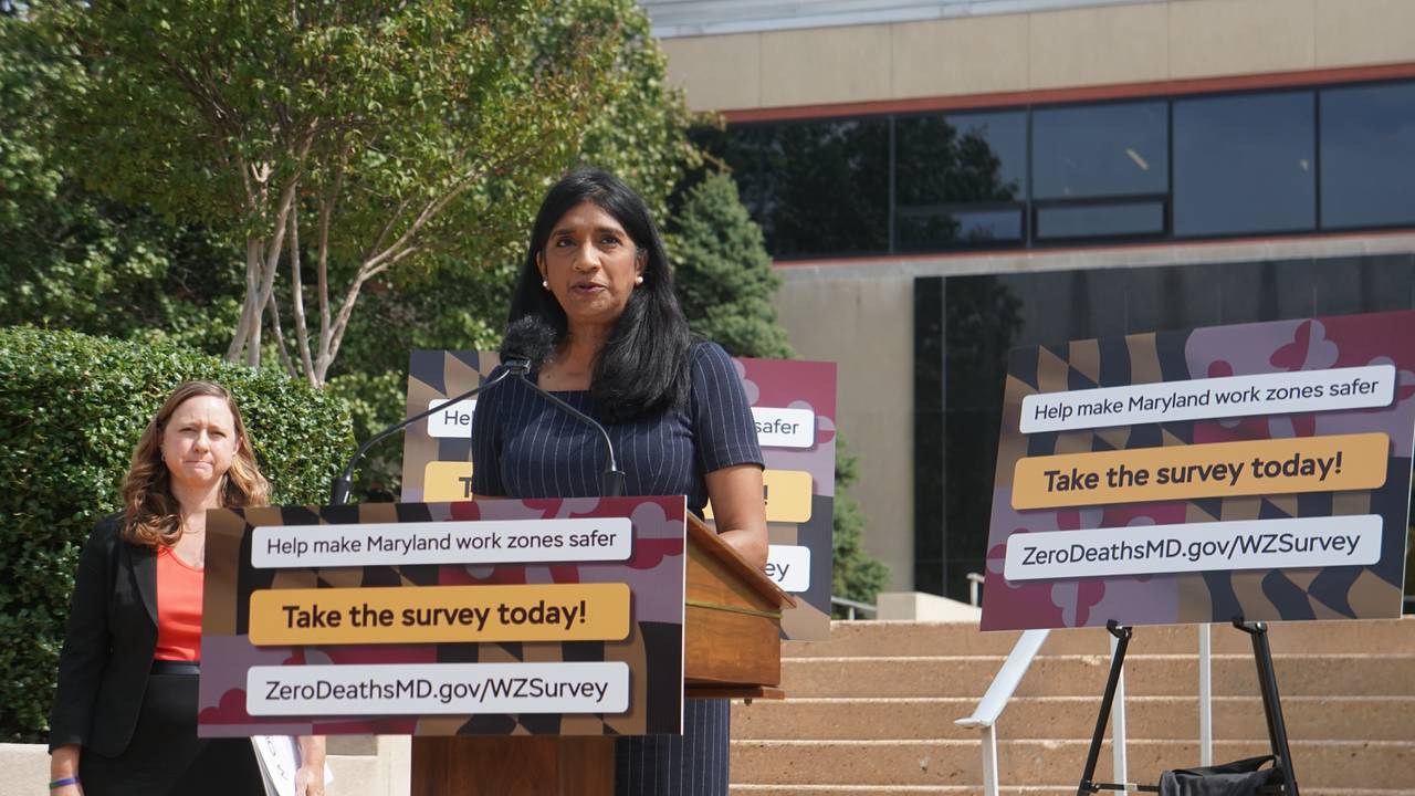 Maryland Lt. Gov. Aruna Miller addresses the media at a podium outside surrounded by signs that encourage drivers to take an online safety survey.