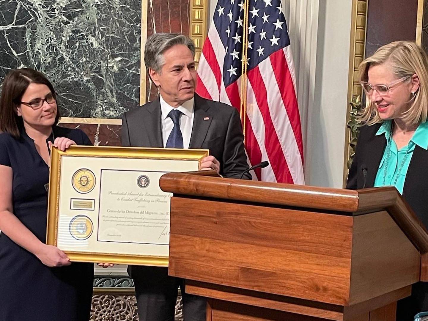 A woman holds a large, framed certificate with a man. Another woman stands at a lectern.