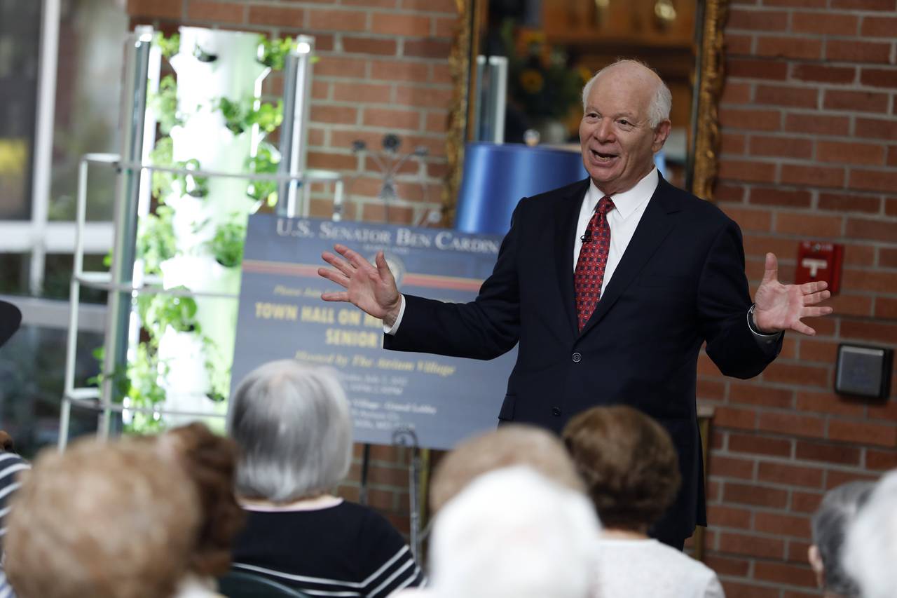 U.S. Sen. Ben Cardin, wearing a dark suit with white shirt and red tie, stands in front of a small crowd with his arms spread wide while speaking.