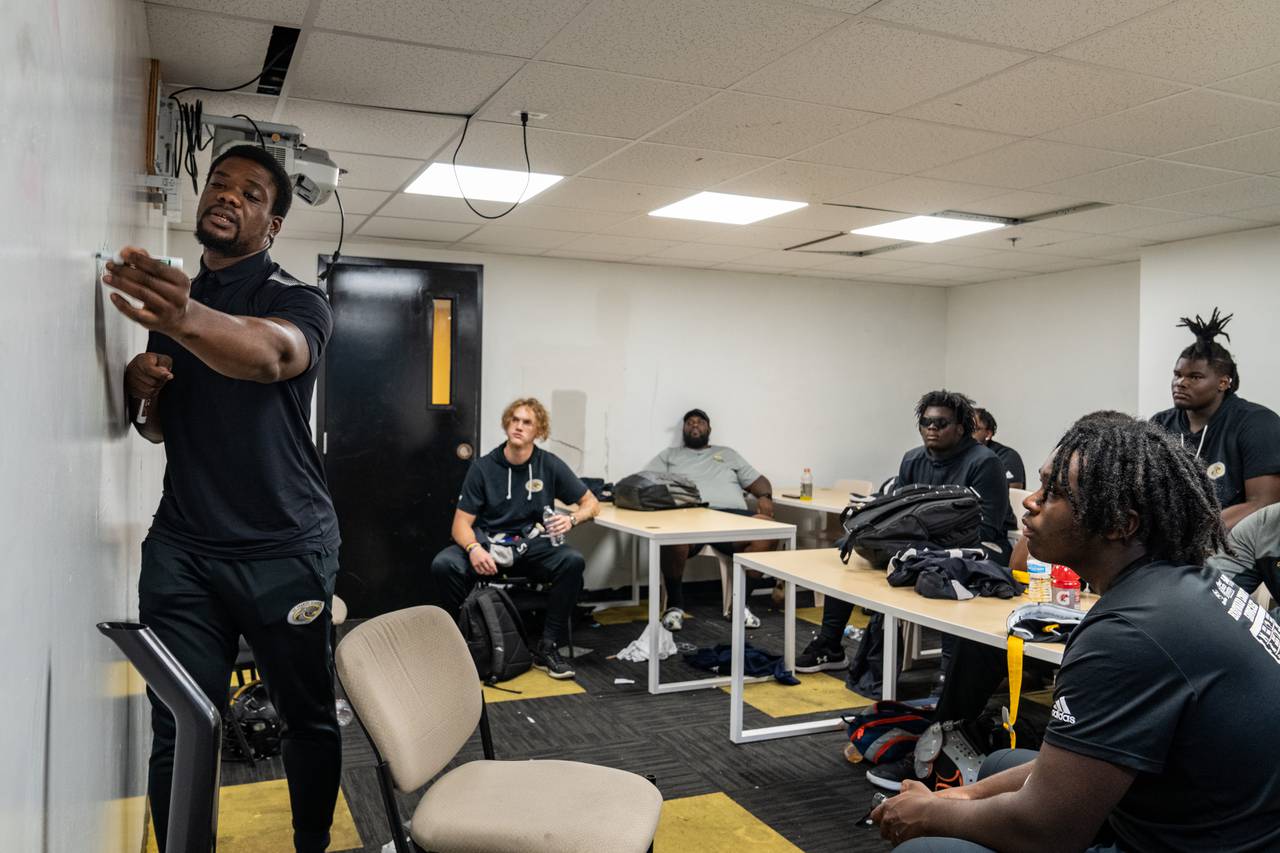Coach Ehijele Ubuane draws on a white board as players on the offensive line sit at tables and listen.