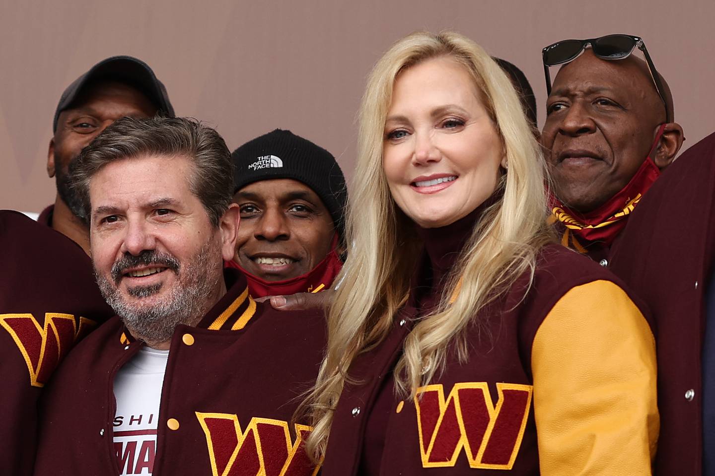 Dan Snyder and his wife Tanya are both wearing burgundy and gold letterman-style Commanders jackets, surrounded by other members of the organization.