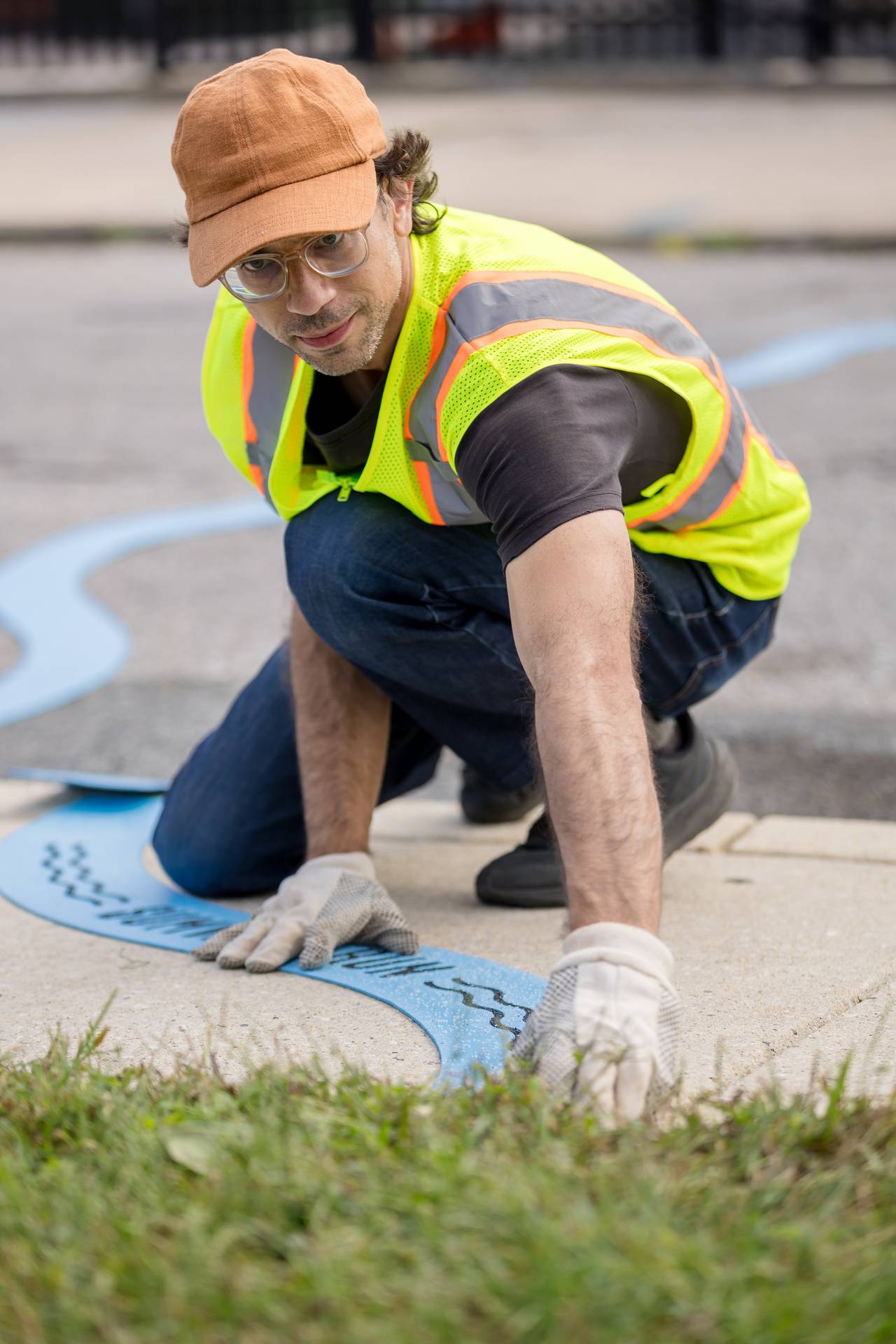 A man wearing a yellow work vest lays blue thermoplastic down on a city sidewalk.