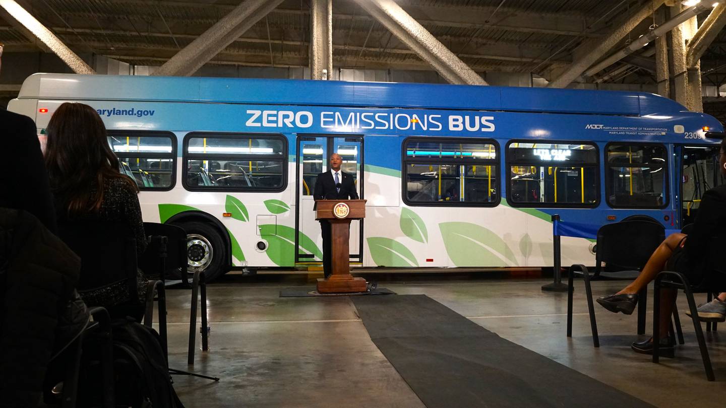 A man stands at a brown podium in front of a blue and white bus that reads "zero emissions bus" on it inside a large bus depot.