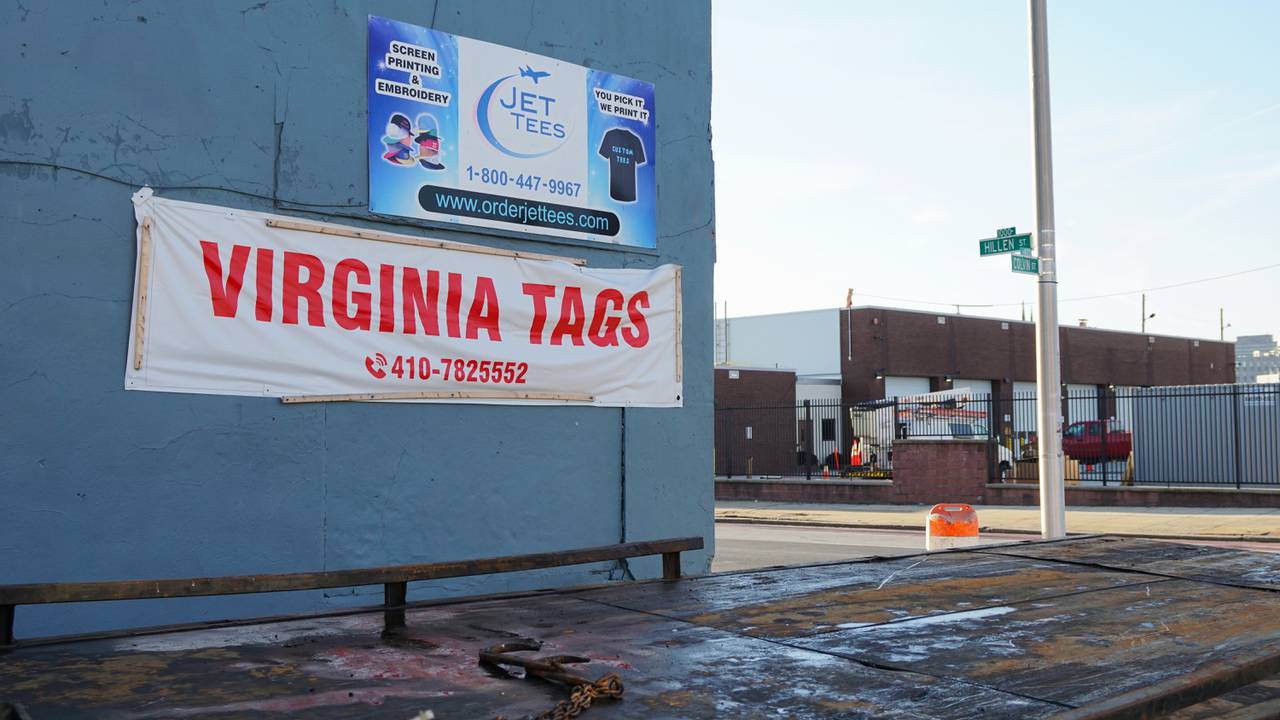 A white and red banner advertising Virginia license plates is hung up on a blue building wall. A truck bed is in the foreground and a city intersection in the background.