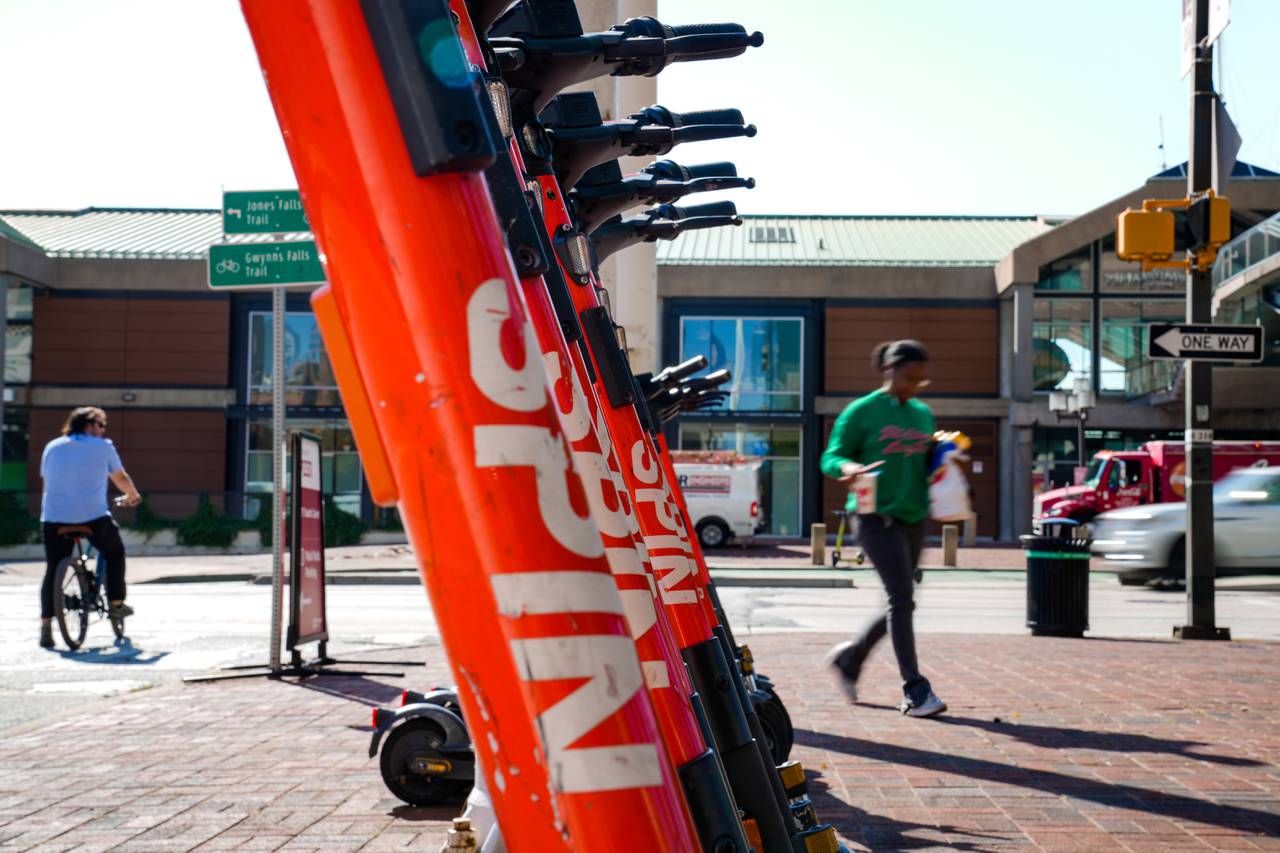 A line of orange Spin scooters divides the frame in half, with a man waiting for a green light on a bike on the left and a woman walking on the right.