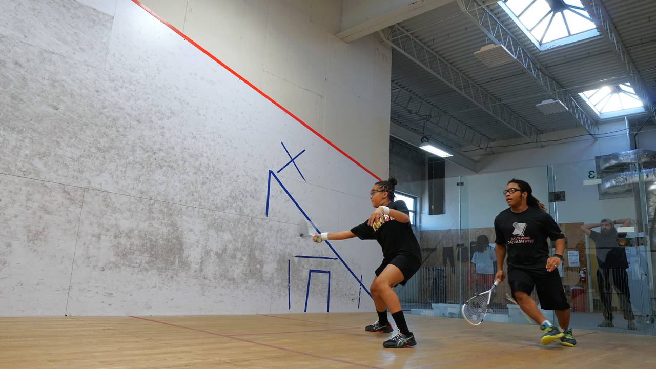 Two teenagers run and swing rackets on a squash court.