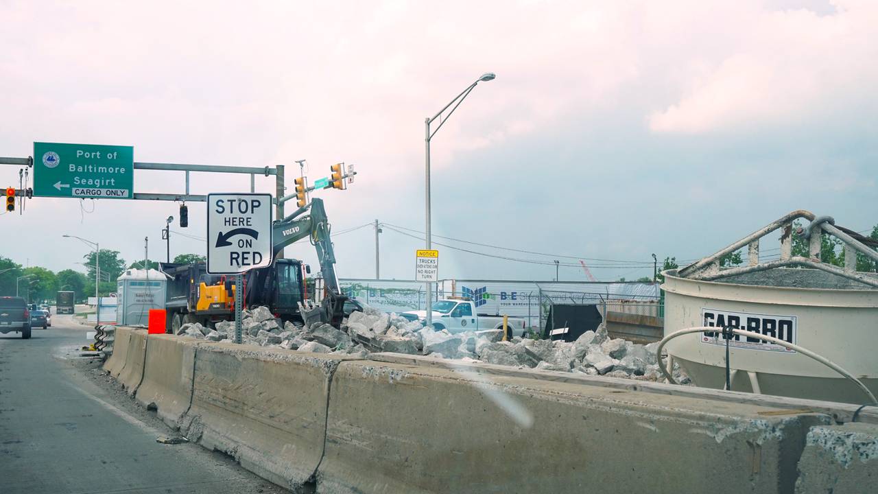 Concrete jersey barriers separate part of a roadway from a construction zone; a crane is removing debris from one section of the site. A green road sign that indicates where to turn for the Port of Baltimore is in the background.