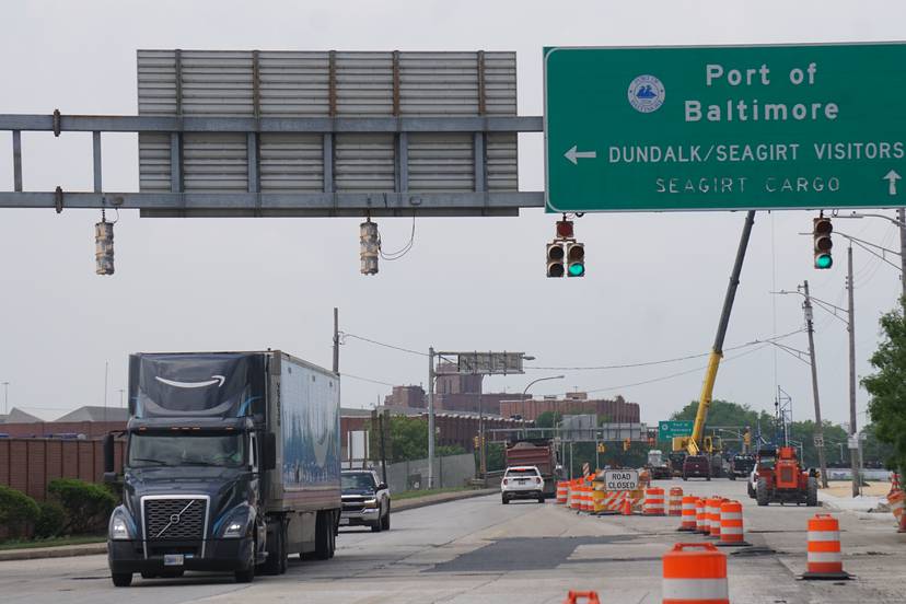 This photo shows a roadway partially closed down with orange construction cones. A truck is driving toward the camera underneath street signs indicating where to turn into a Port of Baltimore terminal.