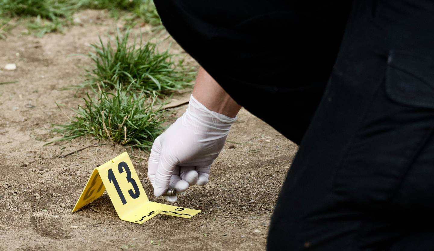 A crouching officer wearing gloves holds a bullet near a yellow evidence marker with the number 13.