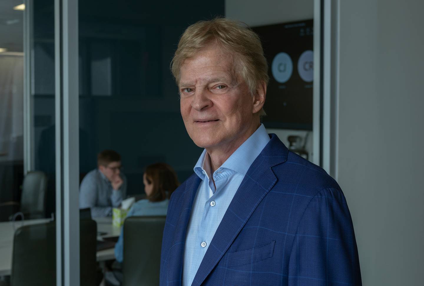 A photo portrait of Stewart Bainum, wearing blue blazer and blue shirt in The Baltimore Banner newsroom, standing in front of a meeting room, with two people meeting behind him.