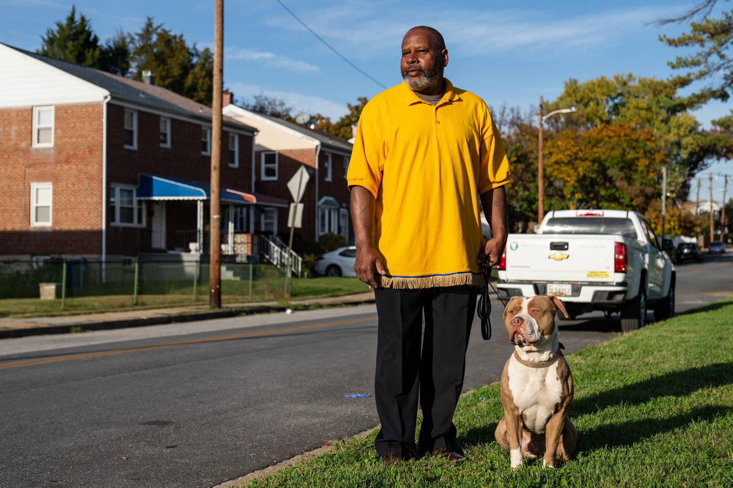 Michael Owens, in a yellow polo, and his white and brown pit bull Champ pose for a portrait on the side of a street, both looking to the left.