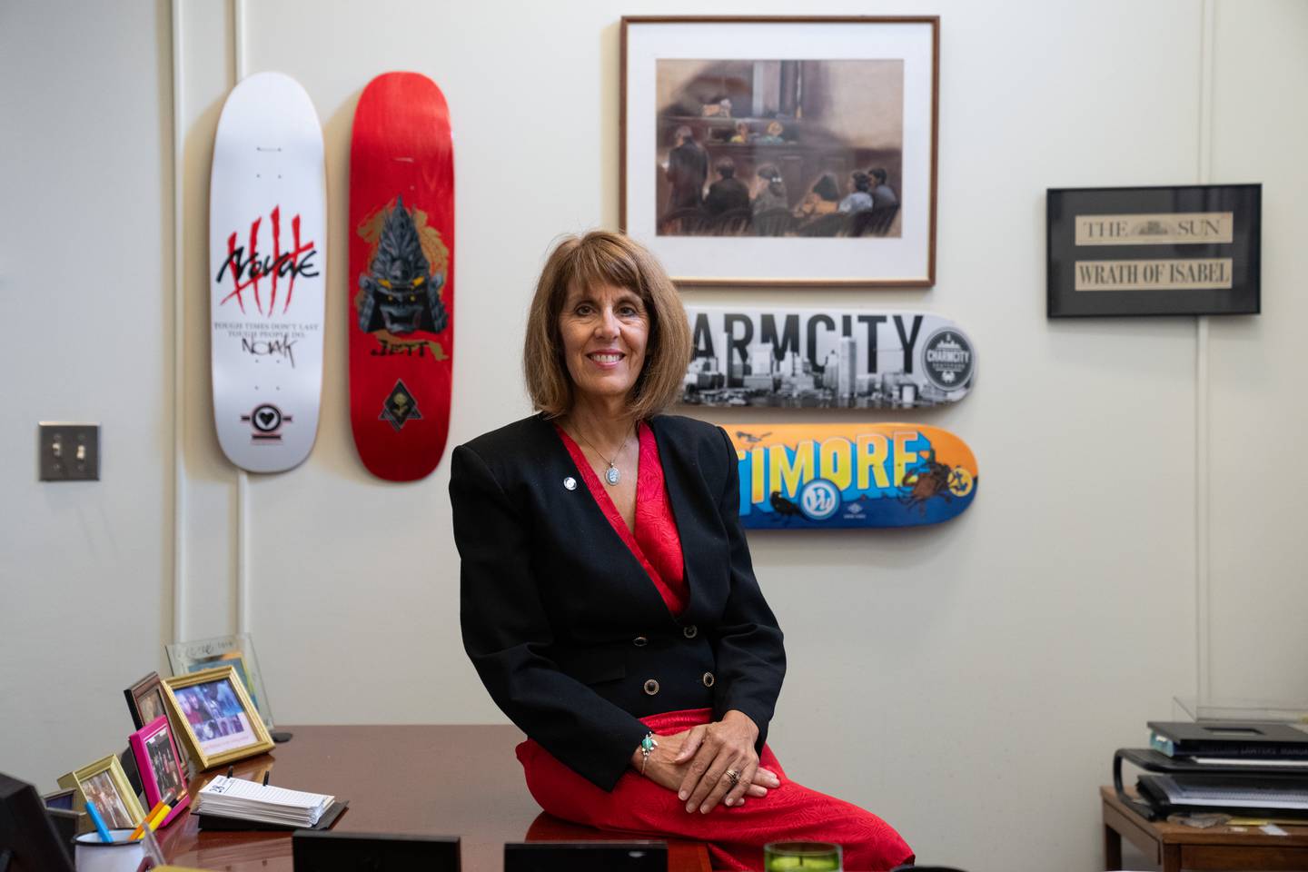 Isabel Cummings poses for a portrait sitting on her desk in her office. Clockwise, the original courtroom sketch of the Jacqueline McLean case she worked on, a Baltimore Sun clipping reading "Wrath of Isabel," a blue and orange "Baltimore" skateboard, a black and white "Charm City" skateboard, one of her son's first pro boards and Brandon Novak's first pro board hang behind her.