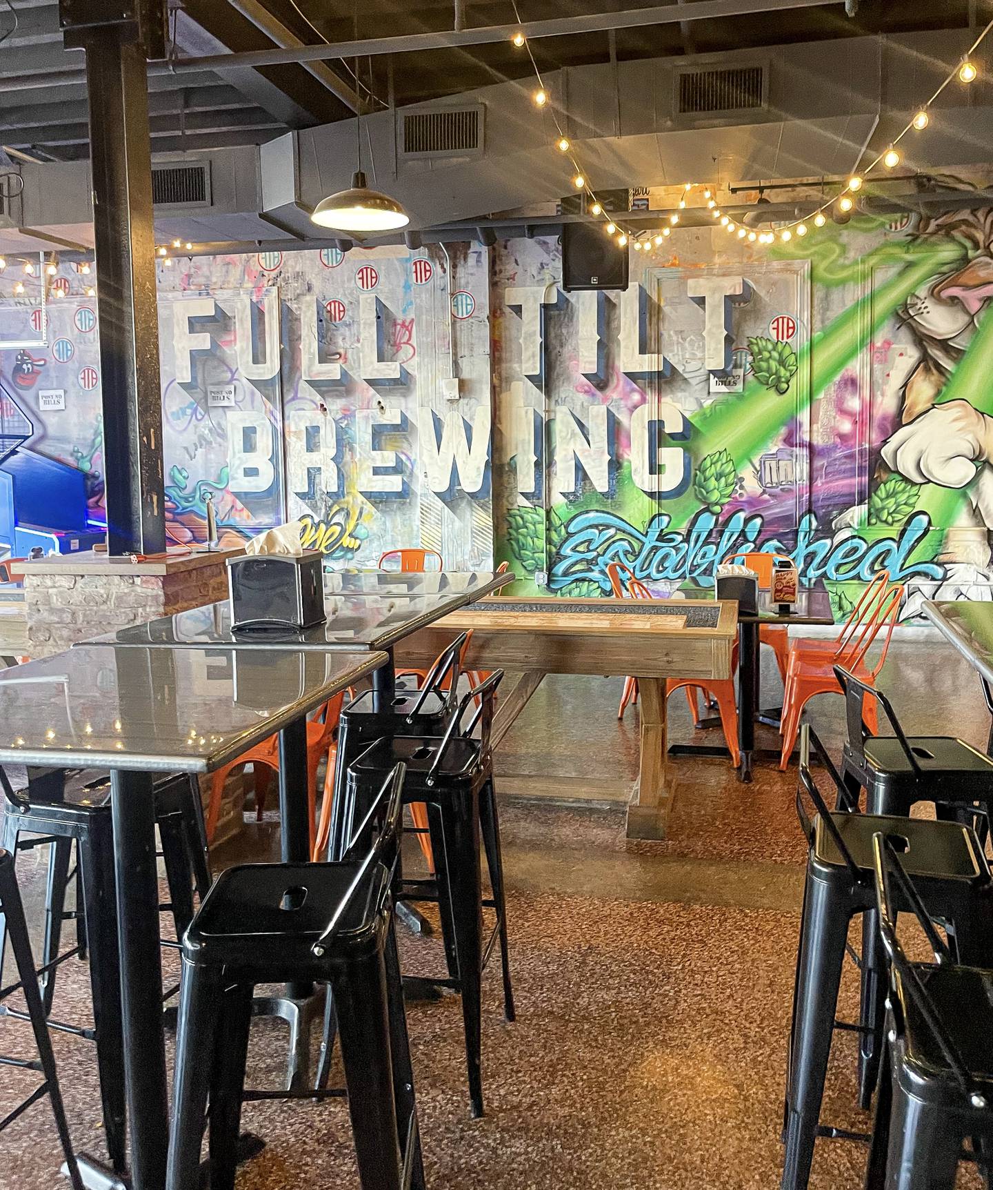 Empty tables and chairs inside Full Tilt Brewing's taproom. A colorful mural in the background reads "Full Tilt Brewing."