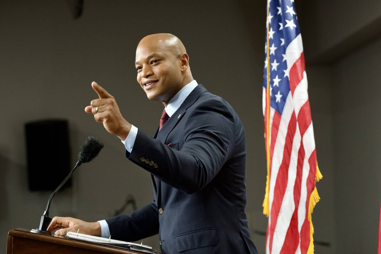 Wes Moore, wearing a dark suit and standing at a lectern, smiles and points with his left index finger. Behind him is a U.S. flag.