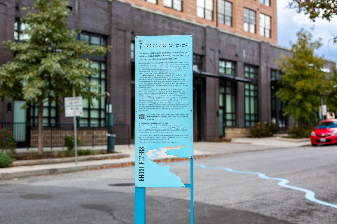 A blue, rectangular road sign covered in black text describes a public art installation on the roadway behind it.
