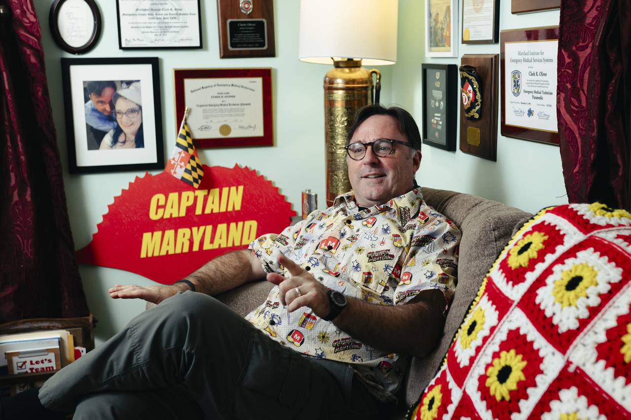 Clark Oliver, also known as Captain Maryland, sits in his living room surrounded by memorabilia from a few years of being superhero, and previously, a few decades of being a firefighter, on Wednesday, Dec. 6, 2023. (Wesley Lapointe/for The Baltimore Banner)