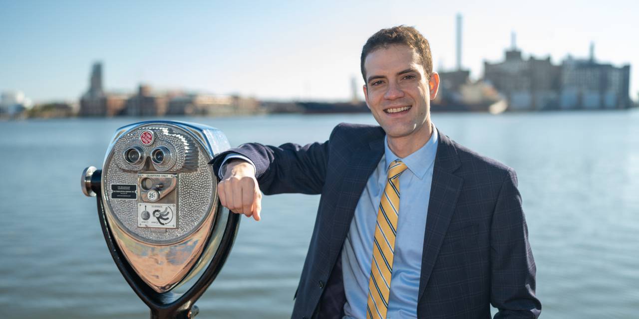 A photo of Liam Davis wearing a dark blue suit, light blue shirt, a gold tie with stripes, in front of the Baltimore harbor. He's leaning on a coin-operated tower viewer.