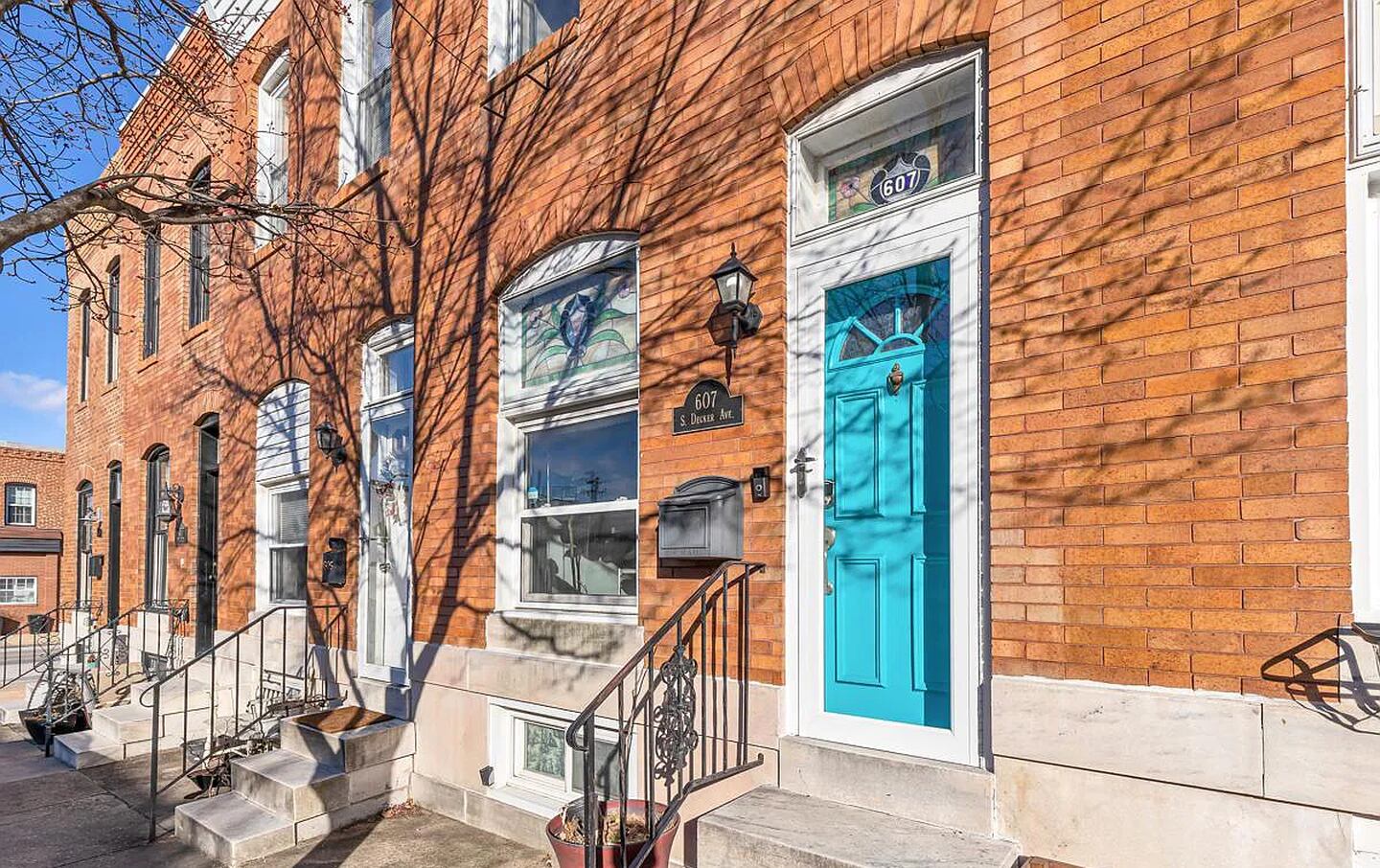 An exterior shot of a brick rowhouse in daylight, with the shadow of a bare tree. The front door to the featured rowhouse is light blue.