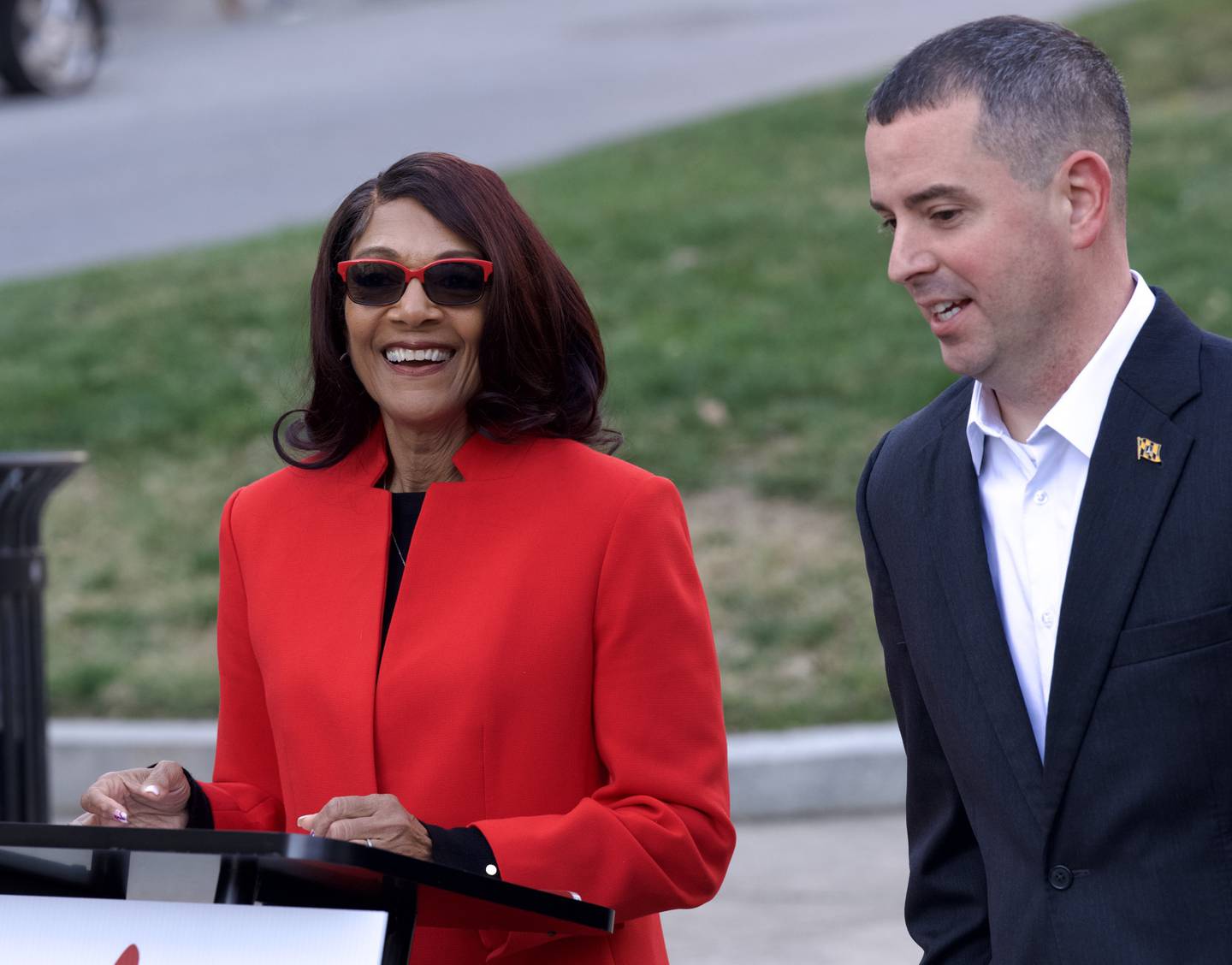 Sheila Dixon stands behind a lectern in a red suit jacket, wearing sunglasses and smiling broadly. To her left, Eric Costello wears a dark suit and white shirt.