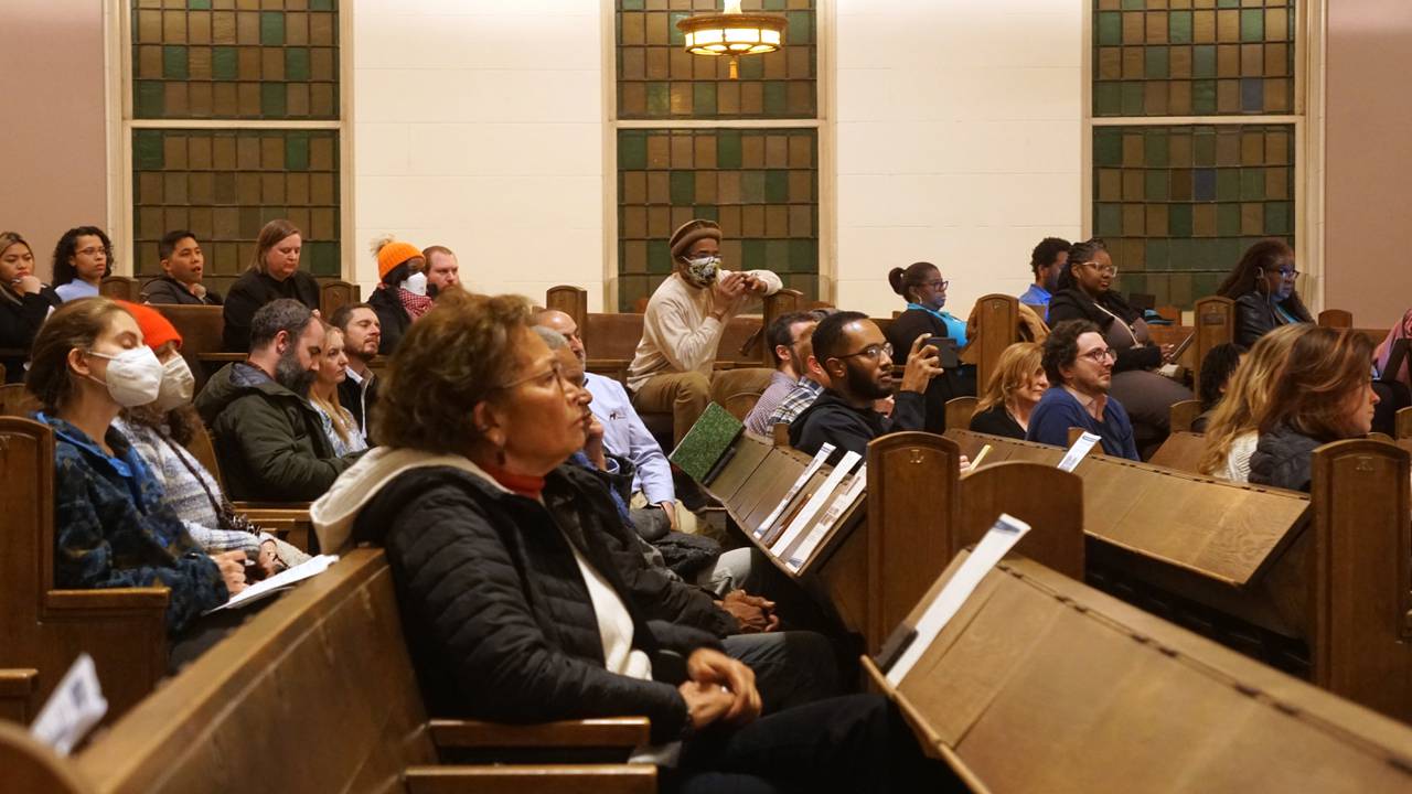 A group of people sitting in synagogue pews looking to the right paying attention to a presentation.