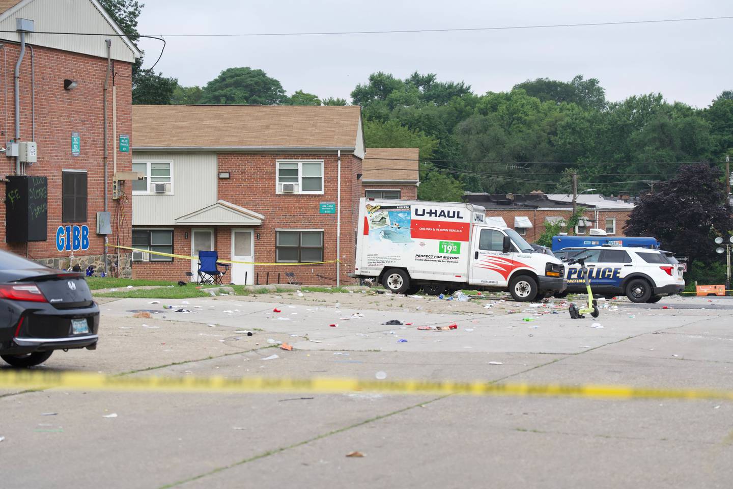 A row of two story brick buildings stands in the background. In the foreground is police tape. A U-Haul truck and police vehicles are visible.