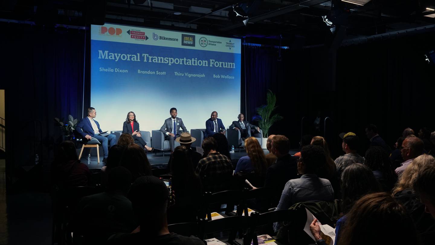 Five people sit on a stage in front of a crowd in a dark room with a projector screen behind them that reads "Mayoral Transportation Forum."