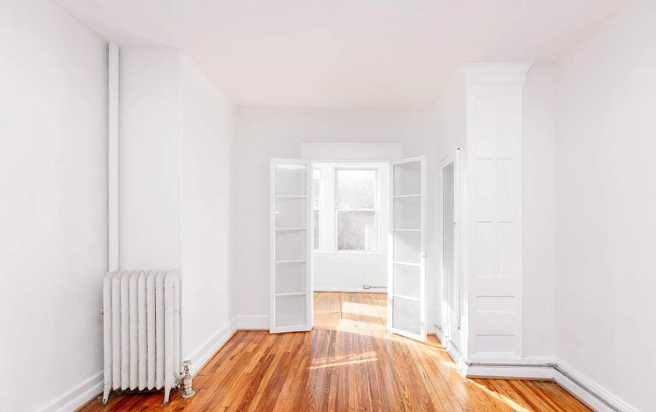 A room with white walls and wooden floors is filled with sunlight. There's a white radiator on the left side of the room and french doors open up into a smaller, brighter room.