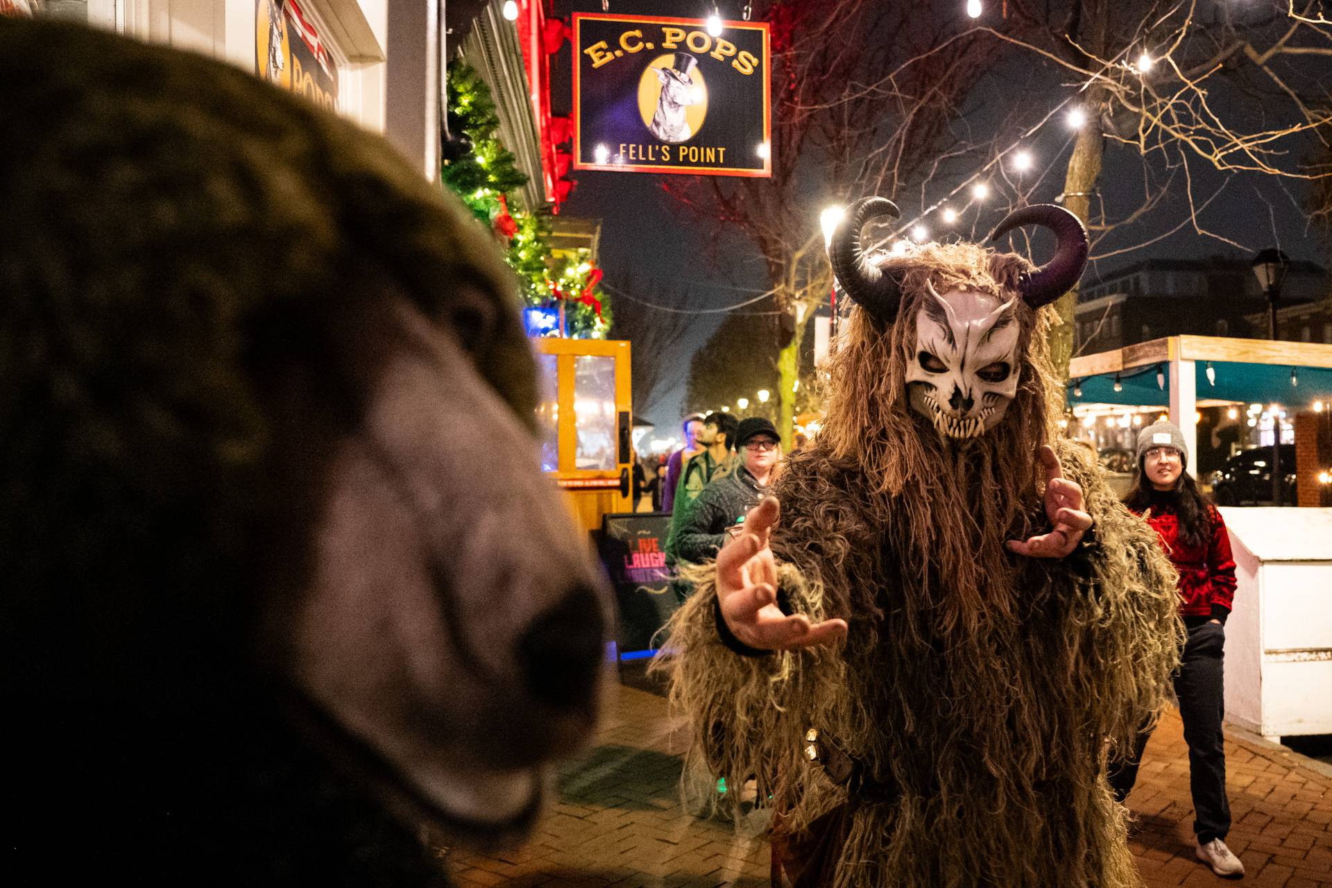 A Krampus wearing a furry moss suit extends his hands toward the camera as he walks by a stuffed bear at E.C. Pops.