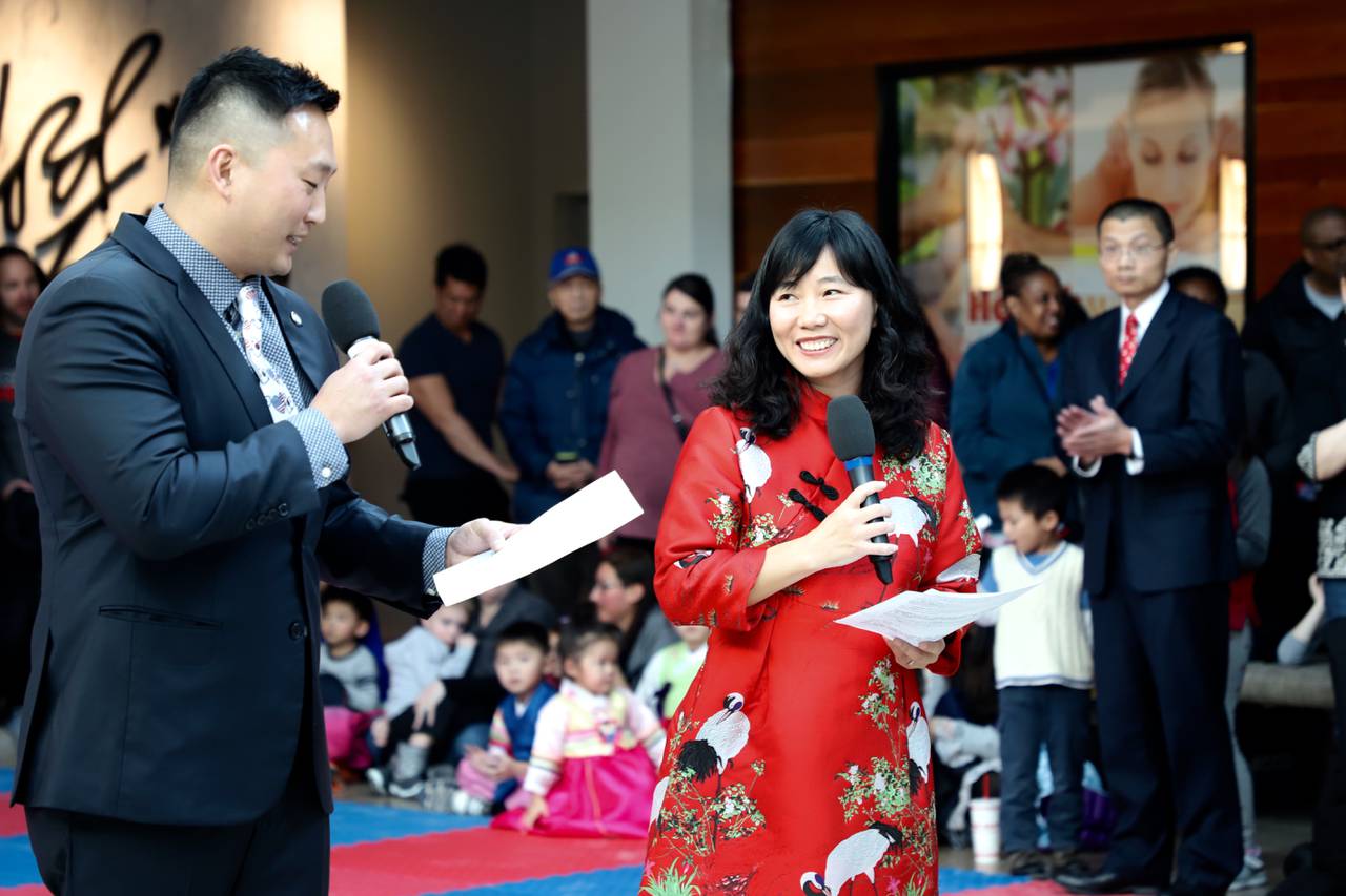 An Asian American woman in a red traditional Chinese dress smiles while holding a microphone. A crowd of adults and children watch behind her.
