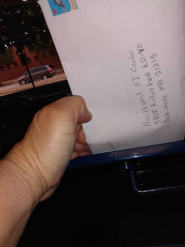 A close up photo of a hand putting an envelope addressed to a state agency into a mailbox.