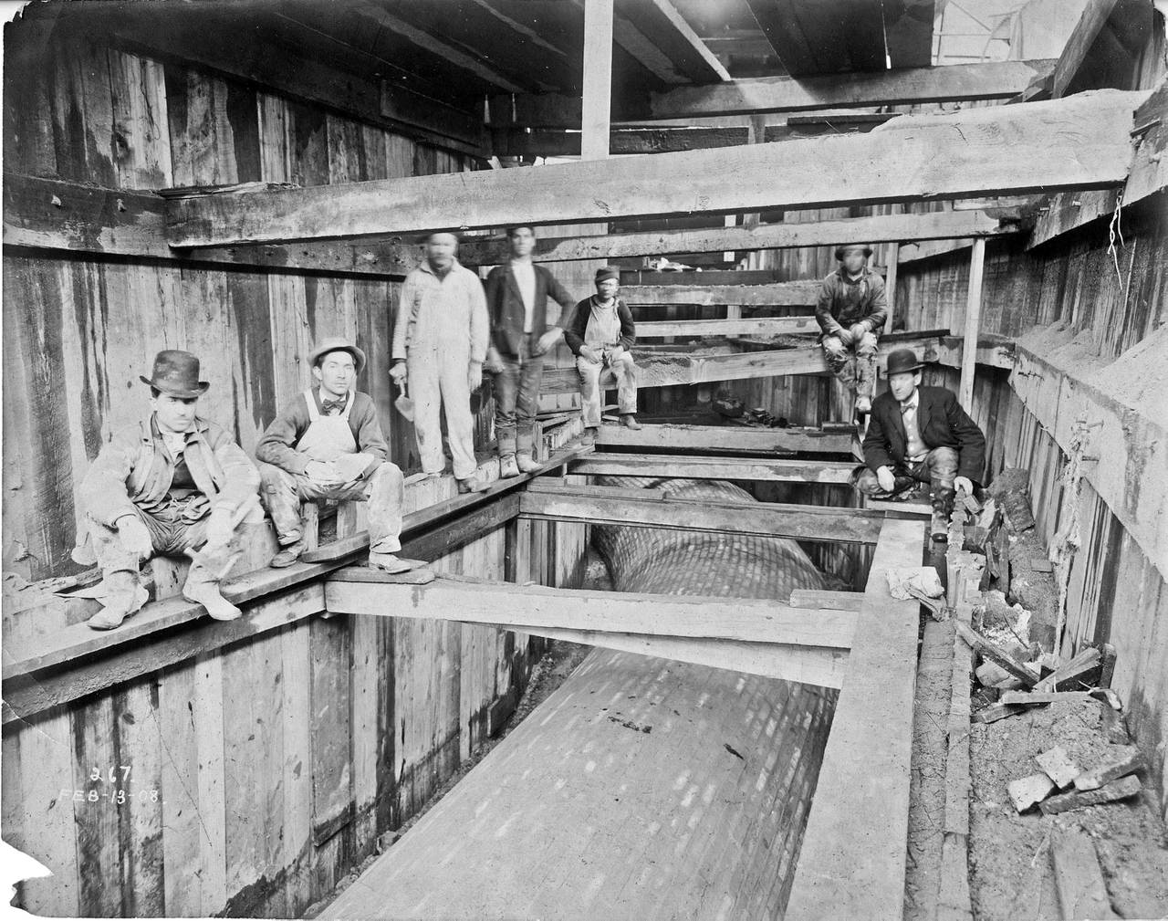 An archived black and white photograph of people sitting on underground scaffolding above a large water pipe.