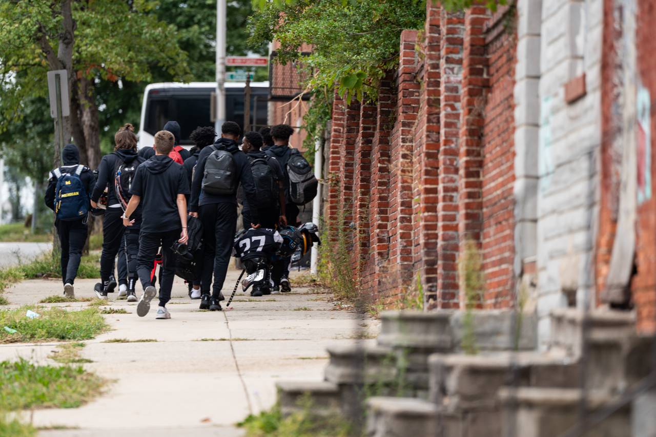 The St. Frances Academy football team walk to the bus, as seen from behind. Brick rowhomes are on the right half of the frame.