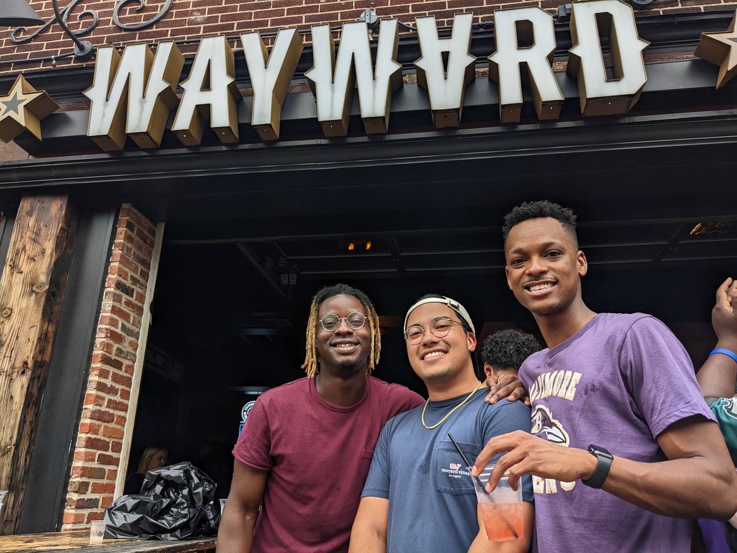 Three men pose for a picture on the outdoor patio of Wayward Bar and Kitchen in Federal Hill. One is wearing a purple Baltimore Ravens shirt.