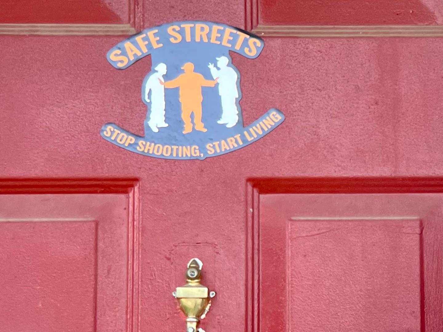 A man in a T-shirt with a white beard stands in front of a red door with a Safe Streets sticker on it.