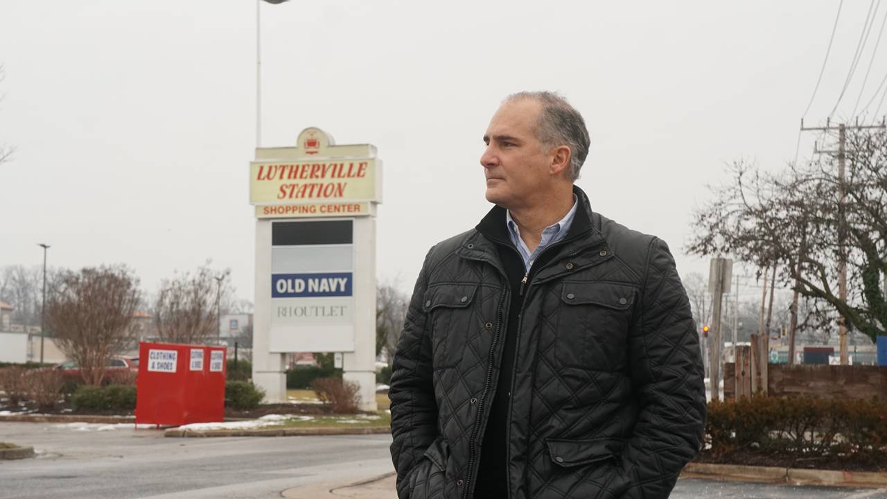 A man wearing a black jacket stands in front of a sign for a strip mall that says Lutherville Station.