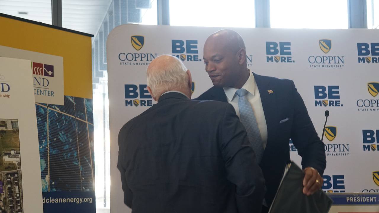 Maryland Gov. Wes Moore, dressed in a suit and light blue tie, shakes hands with U.S. Senator Ben Cardin in front of a white Coppin State University backdrop.