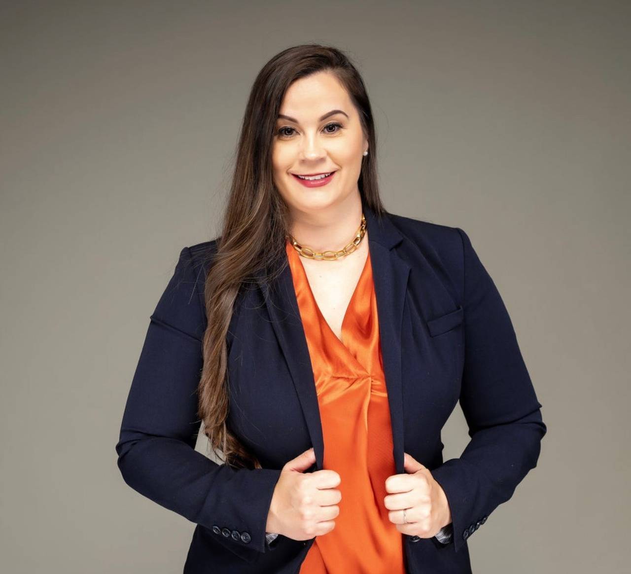 A photo of Margo Bruner-Settles wearing a black suit jacket and orange shirt in a studio with a tan background.