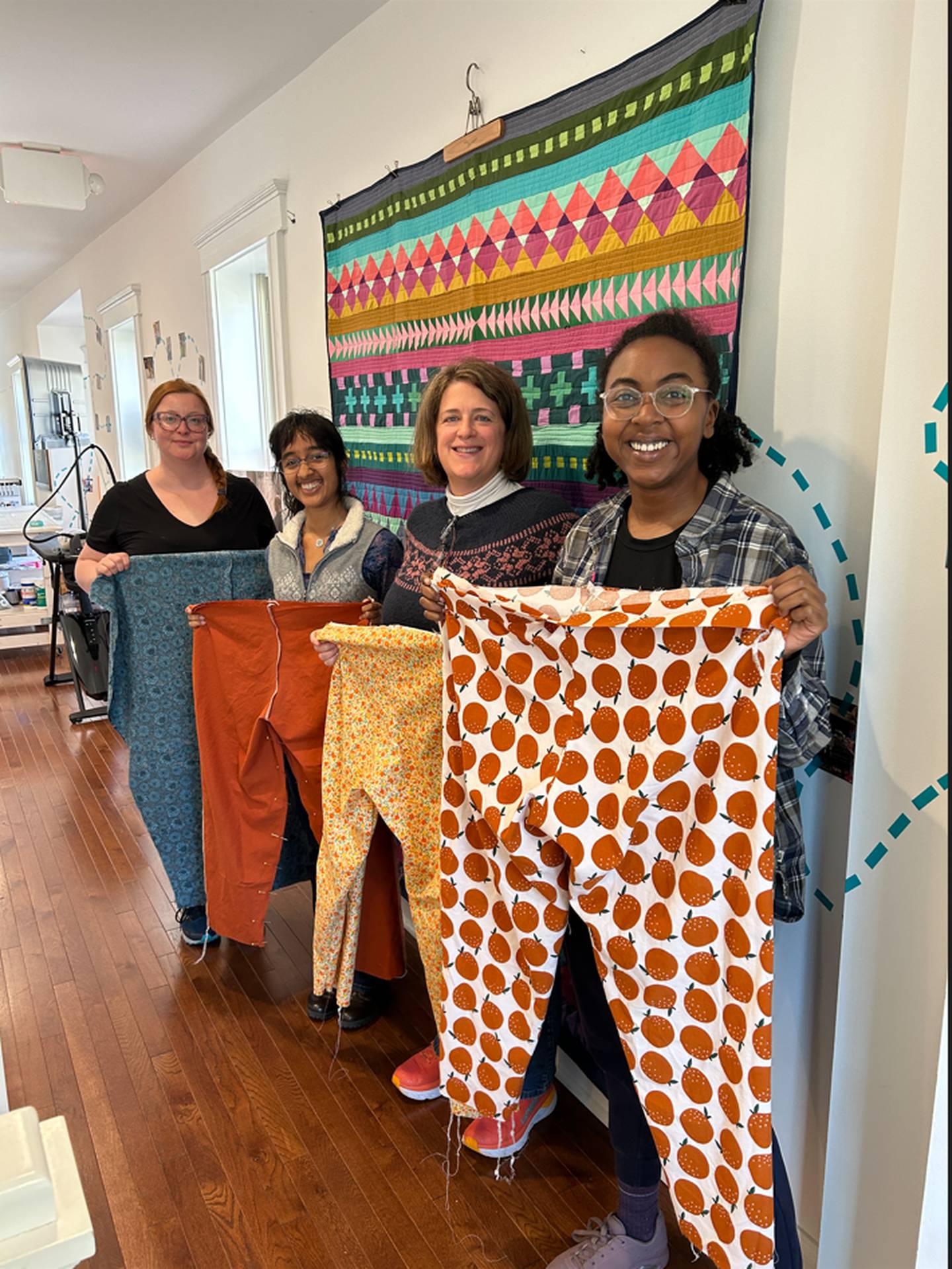 At the end of the Sew Some Garments class, students are holding up their nearly finished trousers. 
From left to right, Cecilia, Neha, Lisa and Imani.