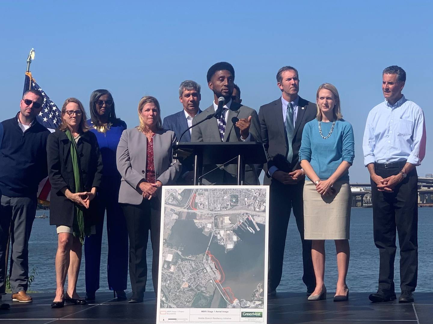 Baltimore Mayor Brandon Scott stands at a lectern with elected officials behind him. In the background is the Middle Branch of the Patapsco River.