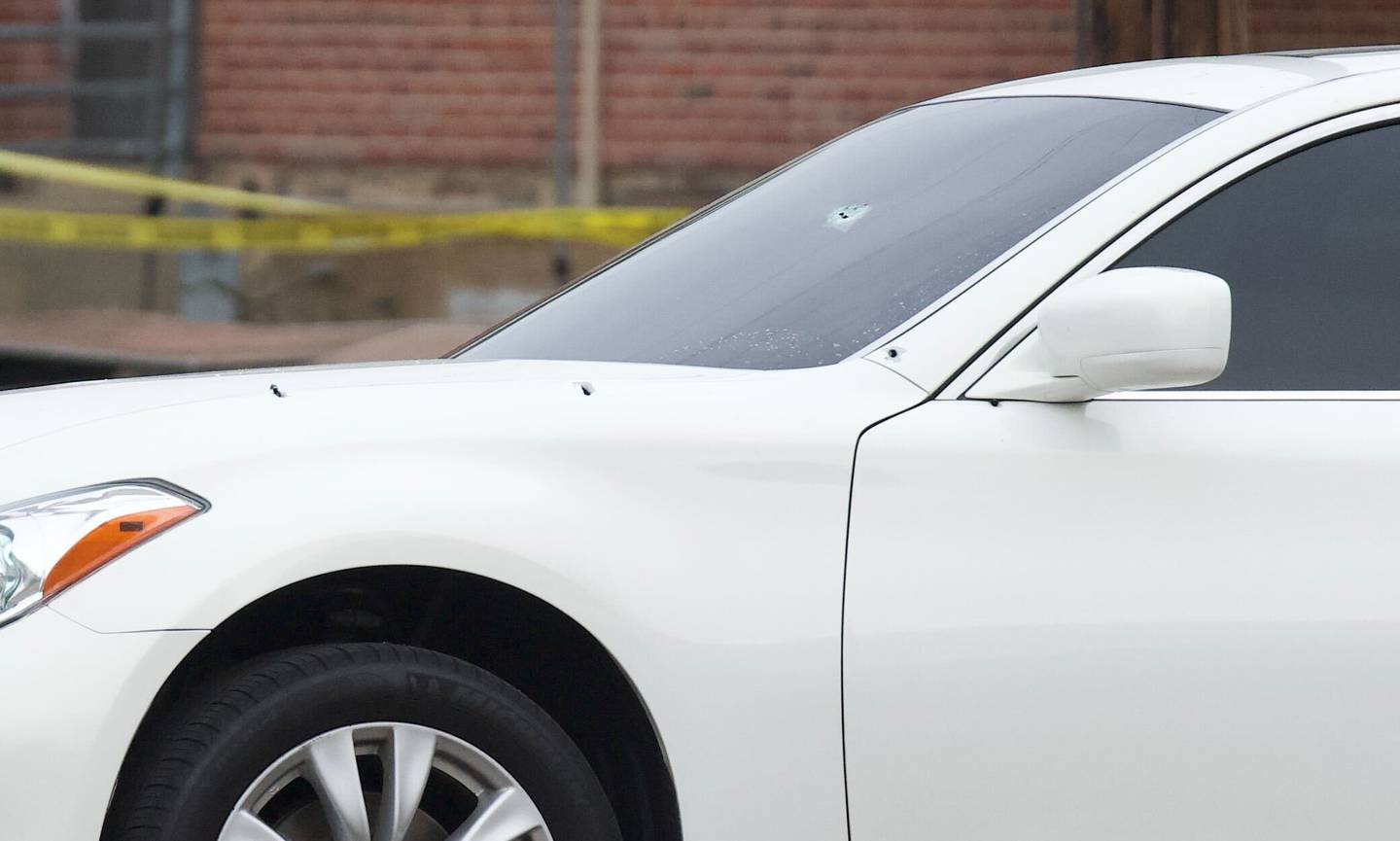 A white car with visible bullet holes near the driver side front door and in the windshield.