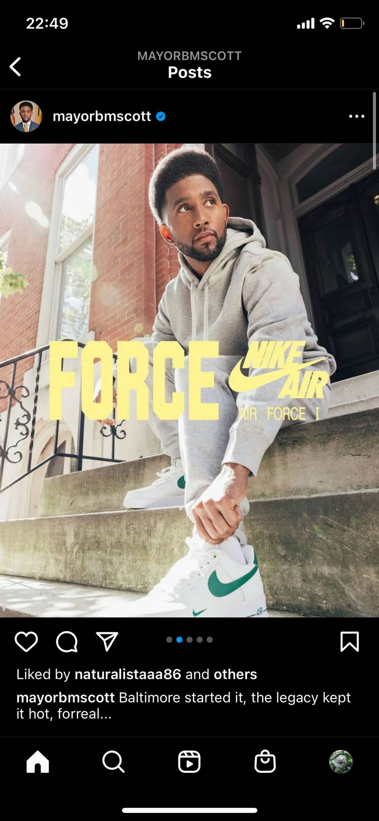 Mayor Brandon Scott sitting on steps in a gray track suit tying the laces on a pair of white Air Force 1 sneakers with a green Nike logo.