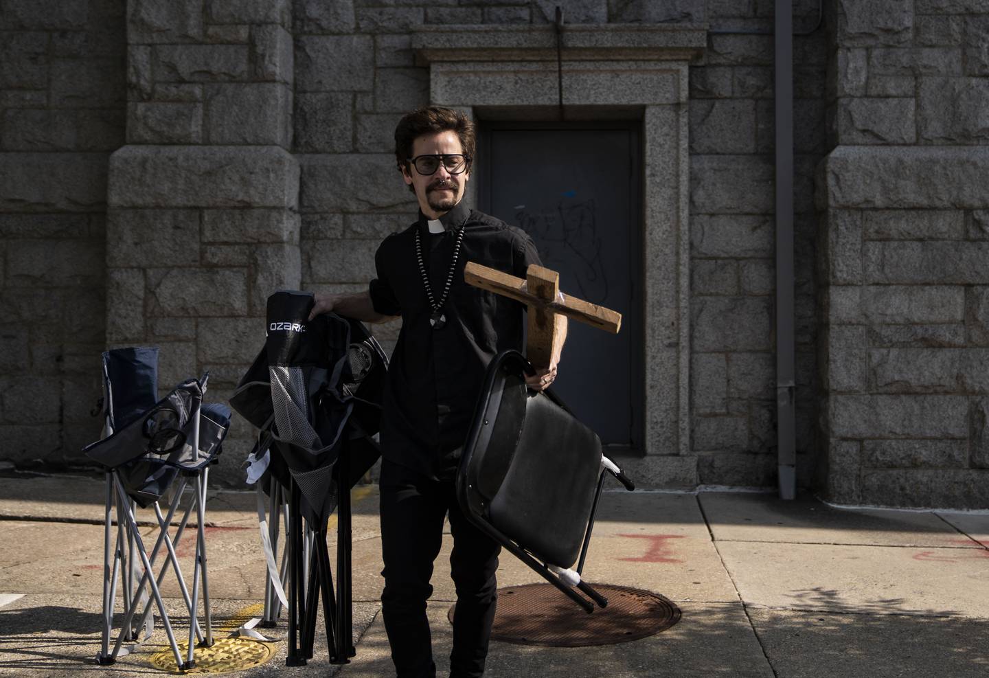 Rev. Elazar Atticus Schoch Zavaletta carries chairs and a cross to their vehicle for the Family Life service at the YNot Lot in Baltimore, Thursday, May 11, 2023.