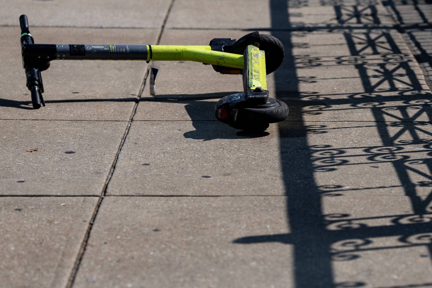 A green scooter lies on the sidewalk on the left side of the frame, as a decorative fence casts a shadow on the right side.