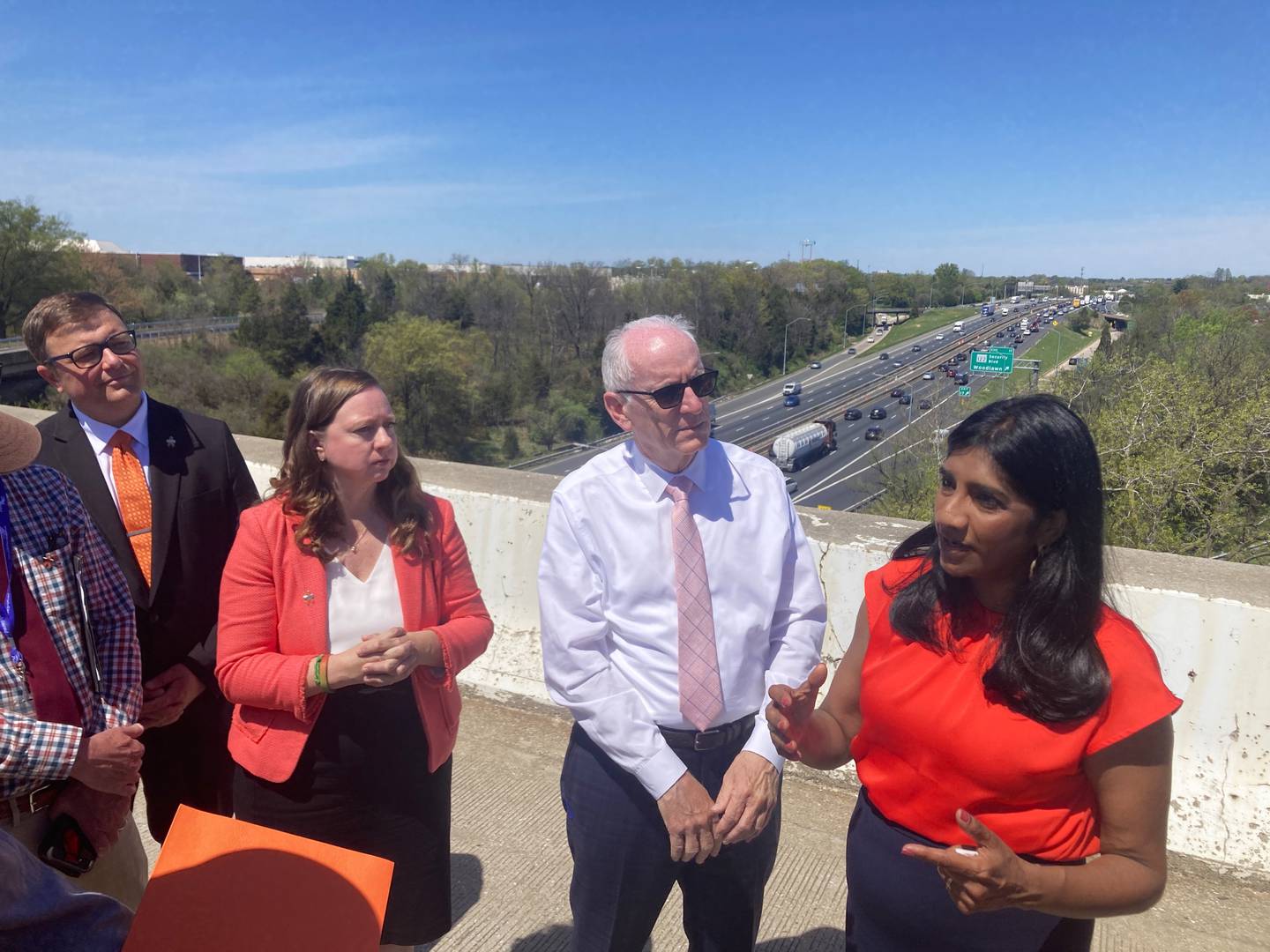 Four people stand and talk on a highway overpass with a large highway in the background.
