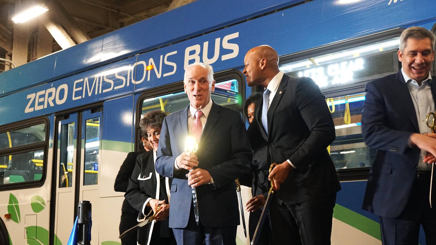 A group of people wearing suits stand in front of a blue and white bus that reads "zero emissions bus" on the side. They are holding large scissors with gold handles they used to cut a blue ribbon.