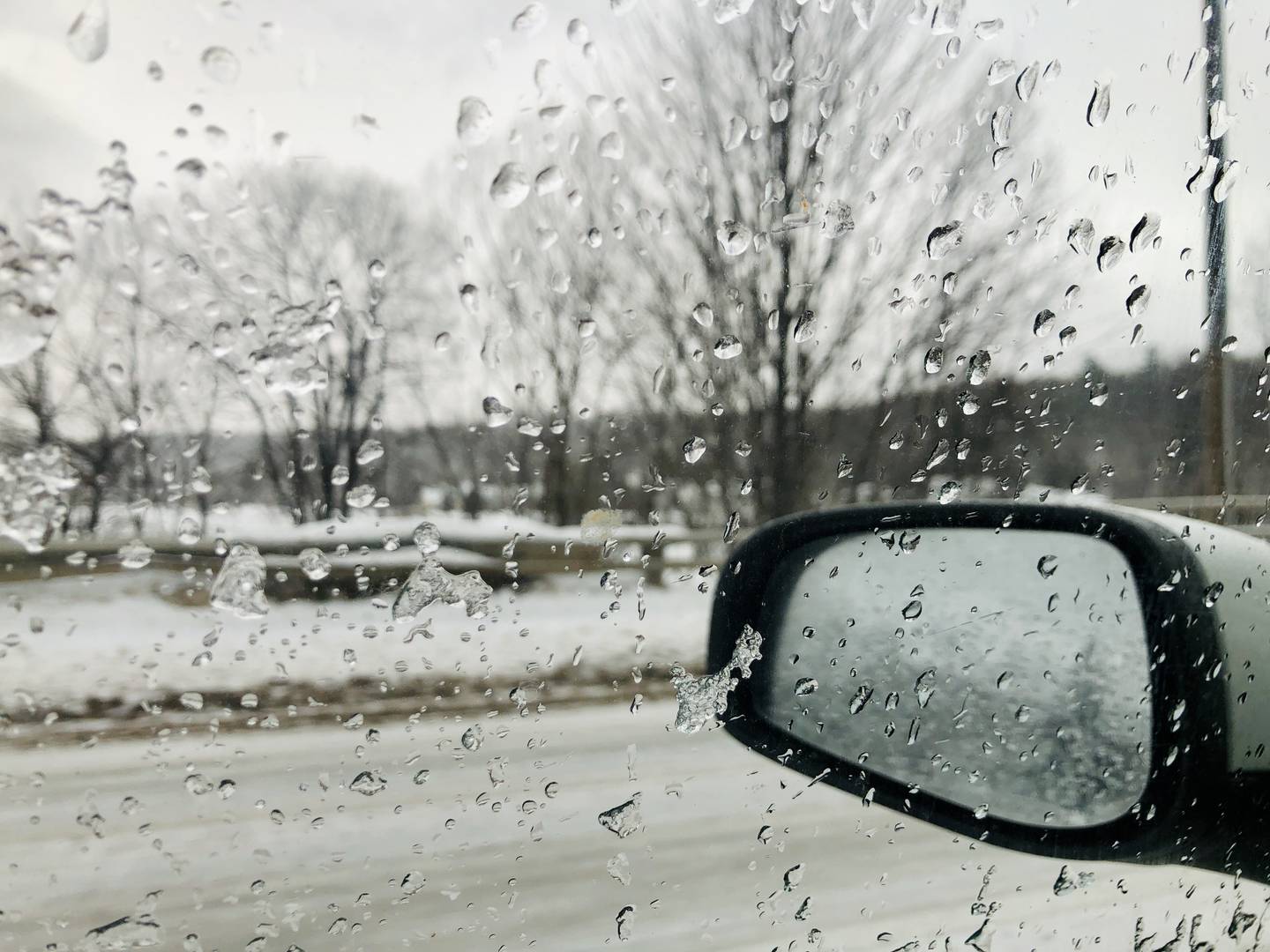 A picture of a vehicle window from the perspective of someone inside the vehicle, looking out. The window is covered in rain and melting frost, with a rearview mirror in the photo and an icy landscape beyond.