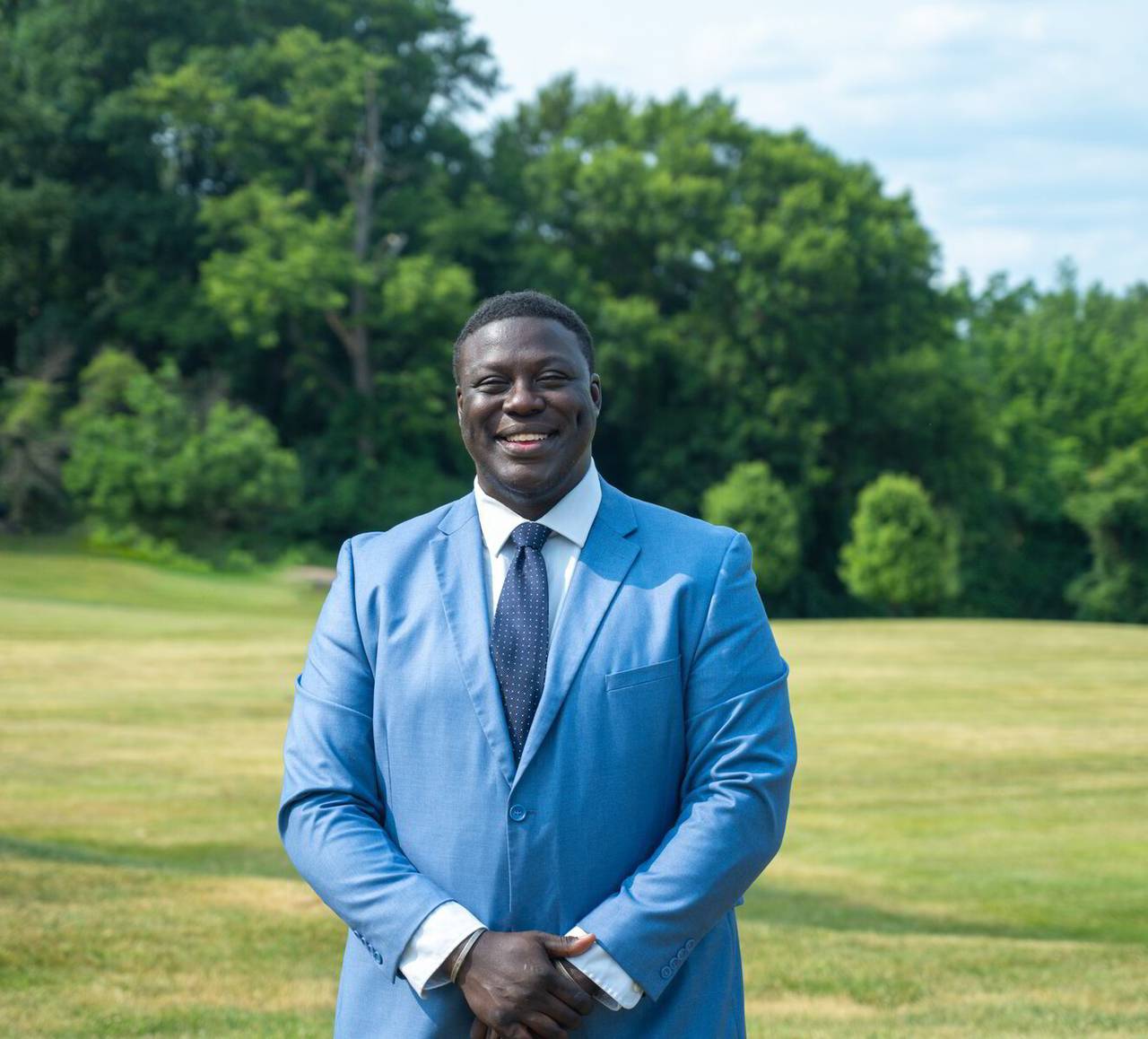 A photo of Steven T. Johnson in a light blue suit, dotted navy tie, and white shirt, with a clear grassy field and trees behind him.